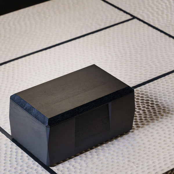 The black lacquered Kist Storage Case with the lid fully closed. The case is sitting on a wooden coffee table with a design that is inspired by Japanese tatami mats.