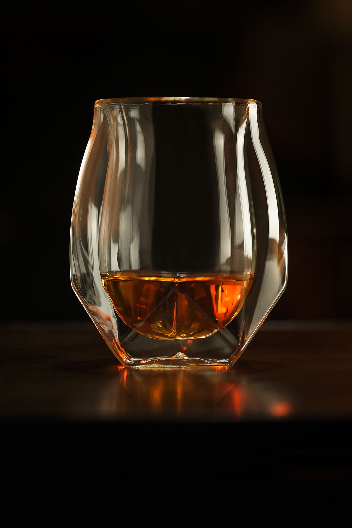 Our unique whisky glass in a darkened room with a sparkling orange hued bourbon whiskey inside. The curvature of the inner and outer walls highlighted against the dark background help illustrate the unique form of this award-winning glass..