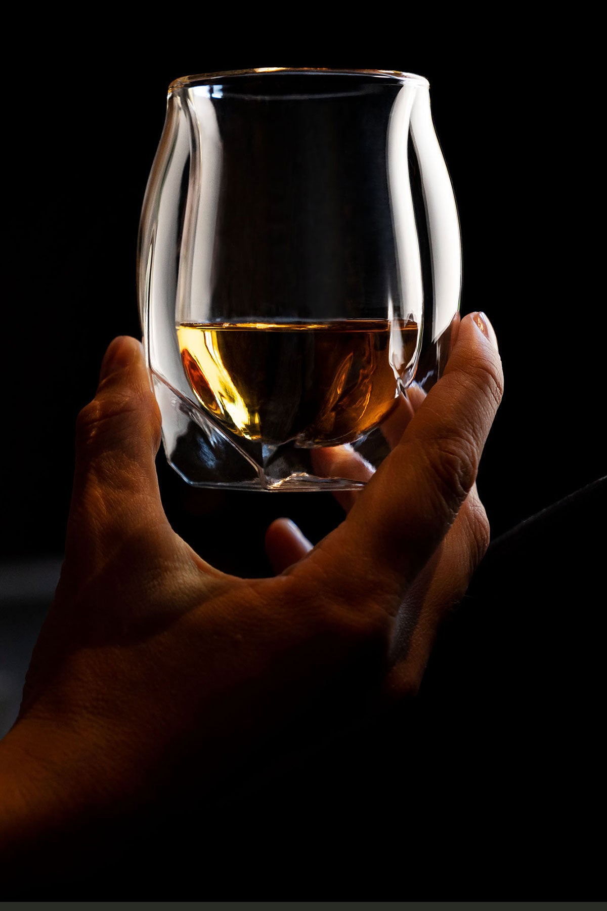 A woman's hand delicately holding tour double-walled whiskey glass by its base, raising it slightly to inspect the honey colored whisky within. The lightness of the glass is implied in the way she is holding the double-walled vessel.