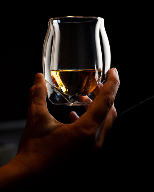 A woman's hand delicately holding tour double-walled whiskey glass by its base, raising it slightly to inspect the honey colored whisky within. The lightness of the glass is implied in the way she is holding the double-walled vessel. 
