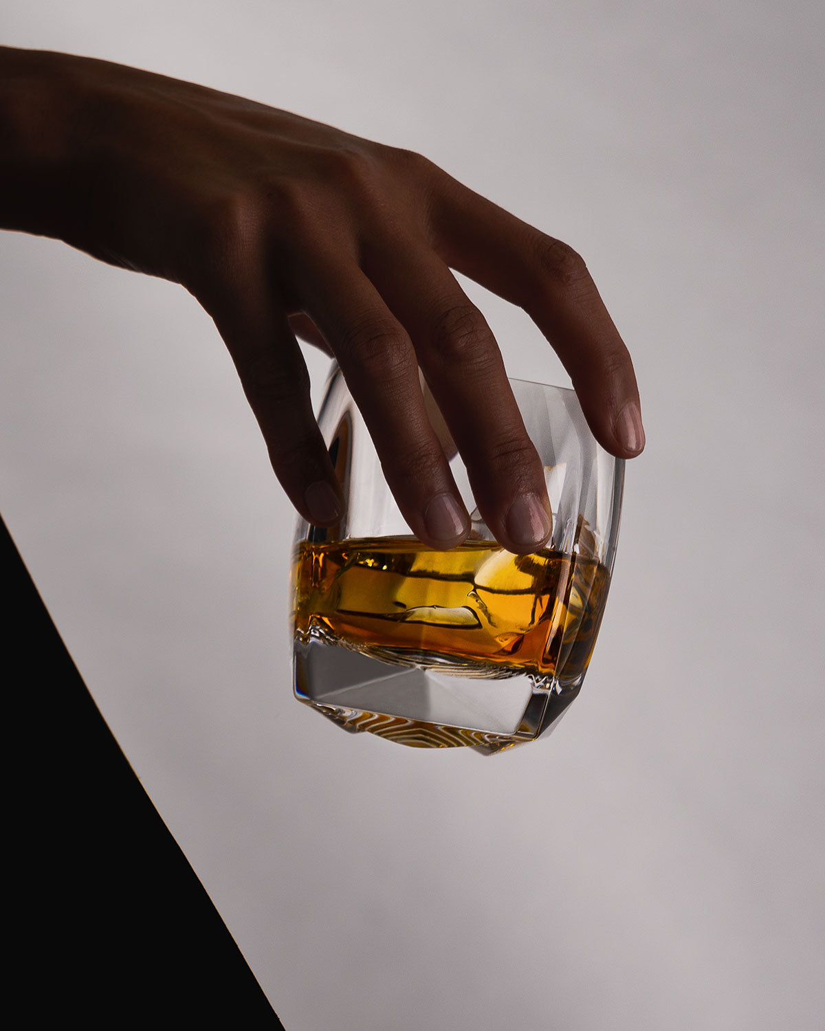 A female hand holding the clear Rauk Heavy Tumbler from near the mouth rim. The faceted surface and three-dimensional base are visible and the weight of the glass is conveyed by how it is being held. 