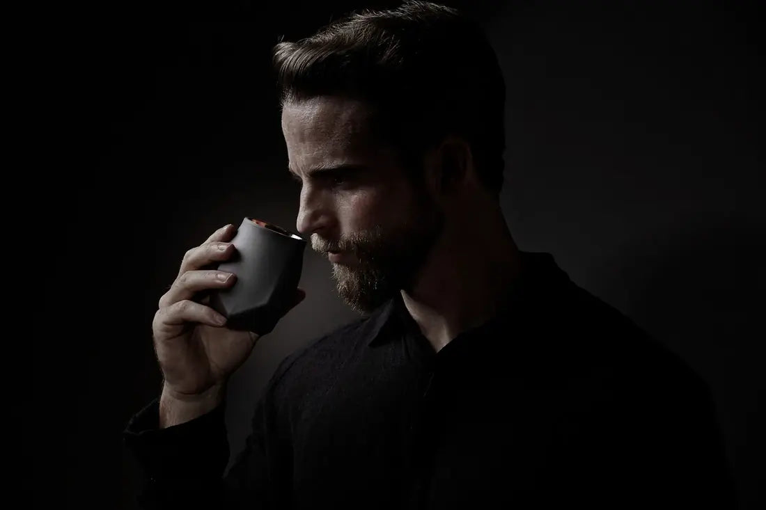 A image of a handsome bearded man against a dark background with a light highlighting his striking features. He hs wearing a black shirt that blends into the background and is holding the Norlan Whisky Glass, VAILD (Black) edition up to his nose to experience the nuanced aromas in his whiskey.