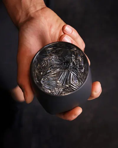 Hand holding the Rauk Heavy Tumbler VAILD (Black) in one hand against a dark background, showing the tumbler without whiskey for a view of the chevron muddling pattern in the bottom of the glass.