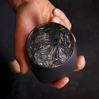 Hand holding the Rauk Heavy Tumbler VAILD (Black) in one hand against a dark background, showing the tumbler without whiskey for a view of the chevron muddling pattern in the bottom of the glass.