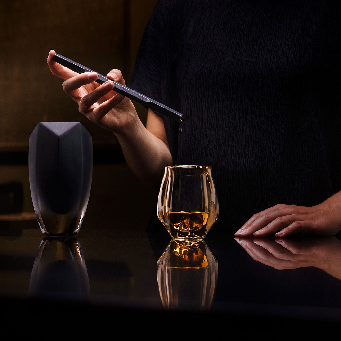 A woman's hand is holding the Drave Water Pipette and adding drops of water to her whisky in the Norlan Whisky Glass. Beside her hand stands the Drave Water Carafe. 