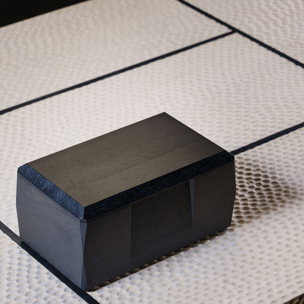 The black lacquered Kist Storage Case with the lid fully closed. The case is sitting on a wooden coffee table with a design that is inspired by Japanese tatami mats. 