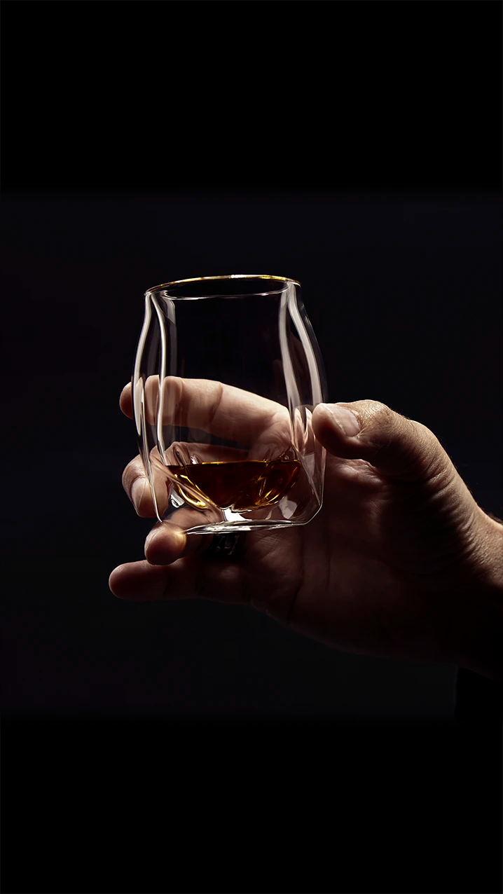 Male hand raising the clear Norlan Whisky Glass to make a toast. The glass is framed by his hand and the background and the whisky within is seen reflected through the mouth rim of the double-walled glass. 