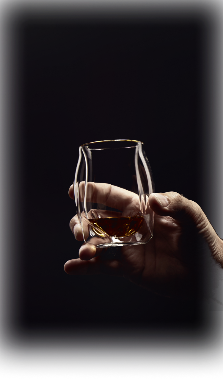 Male hand raising the clear Norlan Whisky Glass to make a toast. The glass is framed by his hand and the background and the whisky within is seen reflected through the mouth rim of the double-walled glass. 