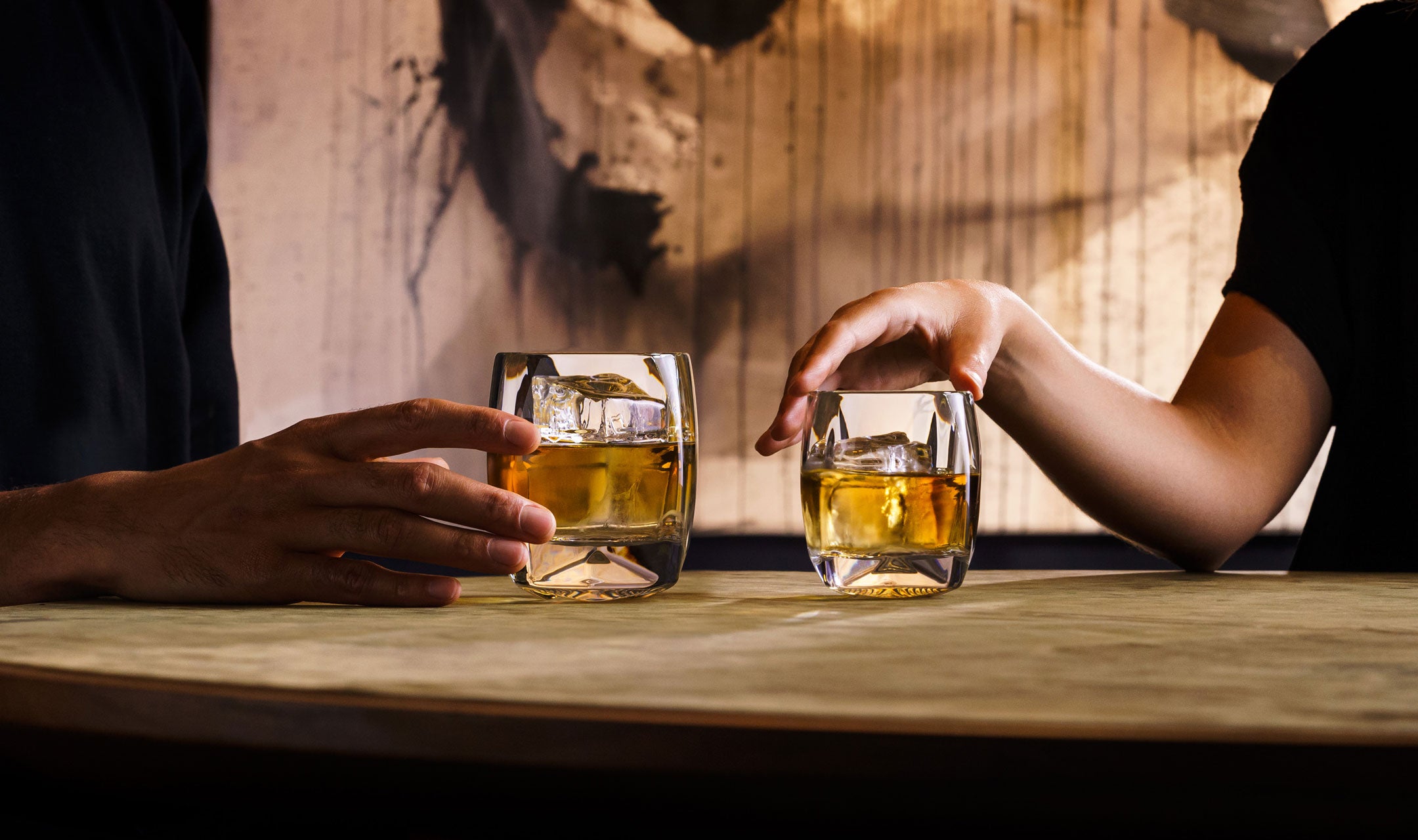 A man and woman are seated at a table. Each is reaching for their Rauk Heavy Tumbler glass. The man has the double old fashioned and the woman has the single old fashioned size. The sizes of the tumblers are therefore easy to see in relation to the hands. 