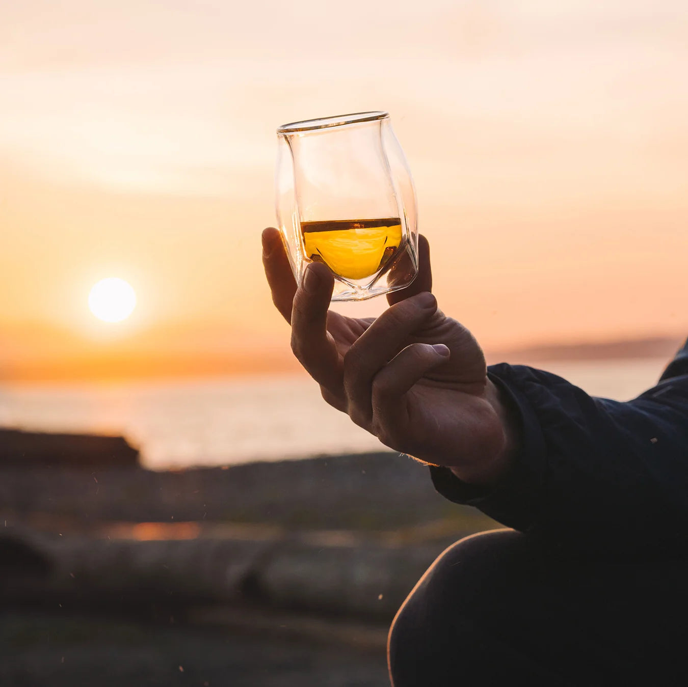 A double-walled whisky glass held aloft on the beach at sunset, the golden glow of bourbon enjoyed outdoors